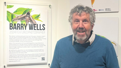 Barry Wells at the 2024 Wildlife Photographer Exhibition. He is wearing a chequered shirt with a blue jumper on top. He is pictured next to a design with his name on it - titled '2024 Featured Photographer'.