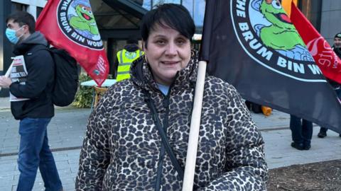 A woman with a leopard-print coat and holding a black flag with a cartoon creature and slogan on it, smiles as she attends a protest. Other protestors, one holding a flag, can be seen behind her.