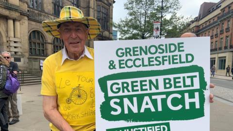 A man is wearing a big yellow checked hat with a bright yellow t-shirt with a bike design. He is holding a large green and white placard saying "green belt snatch" and is standing outside Sheffield town hall.