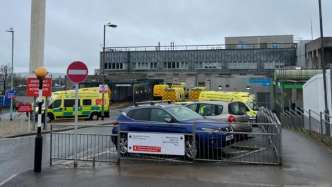 Ambulances and a couple of cars parked outside Derriford Hospital in Plymouth. It is a cloudy and wet day. The hospital building is grey.