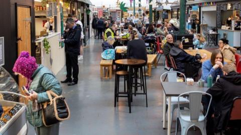 An indoor market lined with stalls on either side and tables and chairs in the centre. The markets is busy with people browsing stalls and eating.