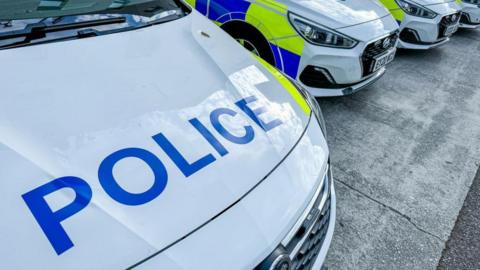 White police cars are parked in a row. The nearest has the word "police" written on the bonnet in large capital letters.