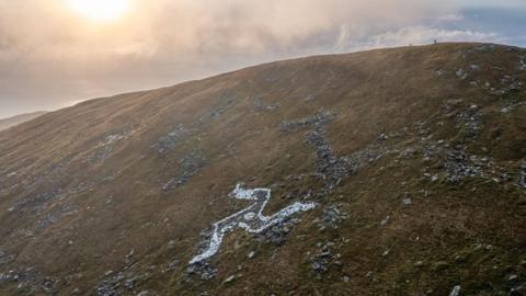 The recently-painted hillside Three Legs of Mann symbol on North Barrule. The landmark's outline and part of its legs are painted white, which stands out against the nearby stone and green hills. There is cloud and a bit of blue sky in the background