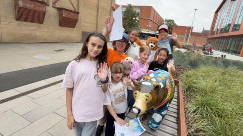A group of children wave beside a colourful painted bear sculpture while holding trail maps in Scunthorpe town centre as part of the We're Going on a Bear Hunt art trail.