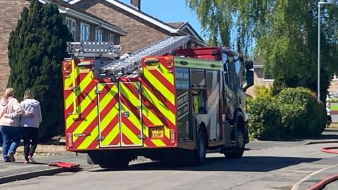 The rear or a fire engine parked outside a row of houses. Two people are walking next to it on the path with their arms around each other.