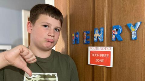A teenage boy stands infront of door with the sign 'tech free zone' attached. He looks unhappy and is pointing downwards with his thumb.
