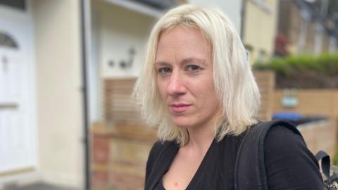 Carly, with shoulder length blonde hair and wearing a black top, is standing in front of a row of houses which are out of focus