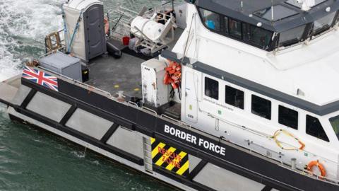 A Border Force vessel leaves the Port of Dover. The close-up photograph shows the equipment on the boat, with a Border Force sign, a Union flag and a "Rescue Zone" sign on the side. The water is choppy and white behind the vessel.