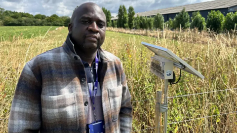 Photograph of Dr Mhango, who is pictured next to one of the monitoring stations. He is wearing a brown and white tartan jacket and has a dark blue university lanyard on. He is standing on the edge of a field which has tall grasses along the edge and a line of trees in the distance. The sky is cloudy, but there is lots of sunshine. The monitoring station consists of a small box attached to a metal stand.