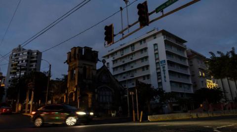 A car drives past dark traffic lights and buildings on a street corner