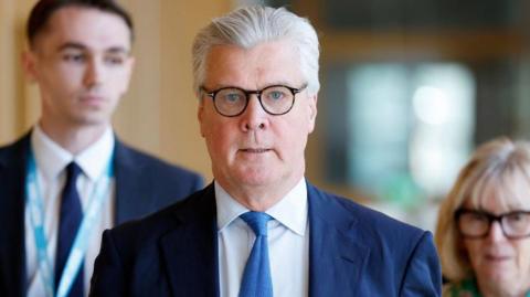Malcolm Offord, who has glasses and grey hair swept back, front on to the camera while wearing a business suit