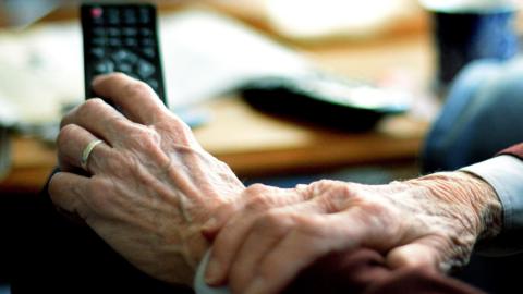 The hands of an anonymous elderly man. He is wearing a wedding ring and hold a remote control.