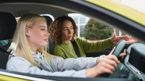 A driver and instructor sit in a car. They are both female and the driver's window is down giving a clearer image of them in the vehicle. The driver has blonde hair and is wearing a grey sweat top and has both hands on the wheel. The instructor has brown curly hair and is in a green top with one hand directing the driver.