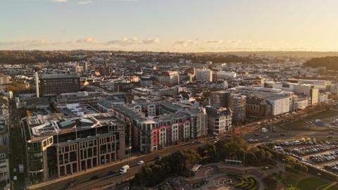 St Helier in evening light viewed from a drone. There is a park and a car park in the foreground and high rise buildings line the main road and stretch back in to the distance.