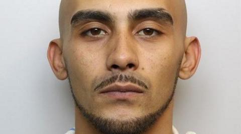 A bald man with a trimmed mustache, beard and thick eyebrows is posing for a mug shot in a police station.