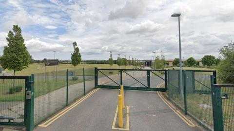 Gated entrance to Sedgefield Community College. The gates and fencing are painted green. Beyond them, a road leads to the main college building. To the sides of the road are schools fields.