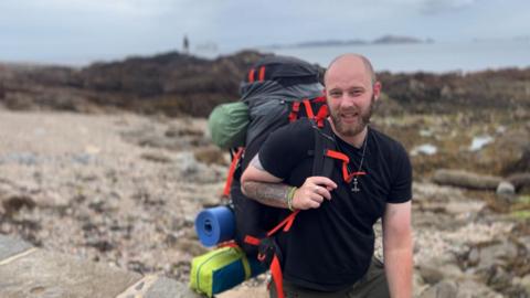 A bald man with a beard in a black T-shirt holds a large backpack on his shoulder, with a rocky beach behind.