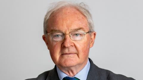 A man with short-receding grey hair stairs into the camera. He wears clear-lensed glasses, a grey suit, blue shirt and navy tie.
