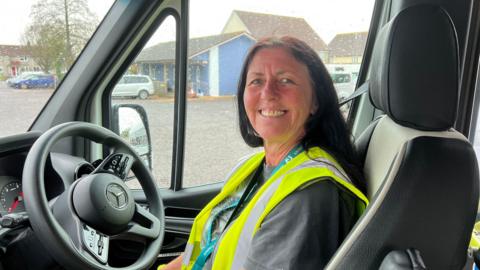 A woman in a high-vis jacket. She is smiling and looking at the camera. She is sitting in the driver's seat of a minibus. In the background there is a bus depot.
