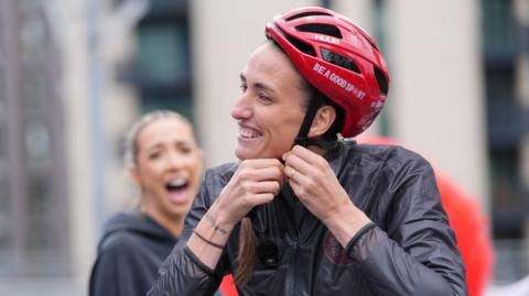 Jill Scott is smiling as she fastens her red helmet. There is some text on the helmet that reads: "Be a good sport." She is wearing a black waterproof jacket and her long brown hair is pulled to the side. GK Barry has her mouth wide open in a big grin behind her but the camera is out of focus.