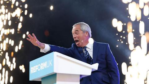 Reform UK leader Nigel Farage on stage at his party's autumn conference. He is waving at the audience at a lectern as fireworks go off around him