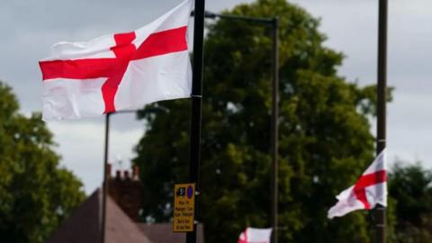 Two England flags hanging from lamp posts with trees in the background