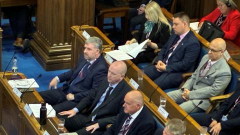 Several men in navy blue and grey suits, shirts and ties are sitting on wooden benches in a council chamber