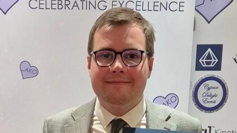 A head and shoulders image of a man, who is wearing glasses, a grey suit, white shirt and dark grey tie. He is looking straight into the camera and he has light, short, brown hair.
