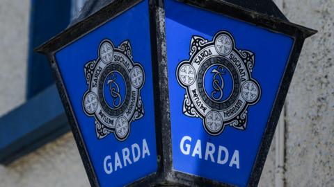  The Garda Siochana logo outside a small Garda station. The walls are white and the logo is displayed on a lantern.