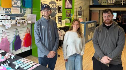 Two men and a woman stand inside a pop-up shop. The space features a wooden floor and walls decorated with framed pictures and merchandise. Clothing and accessories are displayed throughout.