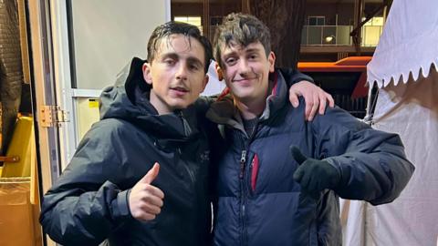 Josh Bennett is posing for a photo alongside Timothée Chalamet. They both are giving a thumbs up to the camera.