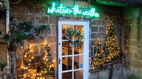 Christmas lights around a door in Broadway, Worcestershire. A lit sign above the door reads: "Whatever the Weather"