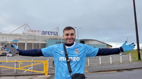 man with brown hair stood outside the Coventry Building Society Arena near a yellow car park barrier with his arms outstretched. The man is wearing a blue replica Coventry City football shirt with a long-sleeved navy t-shirt underneath with blue gloves with navy fingertips.