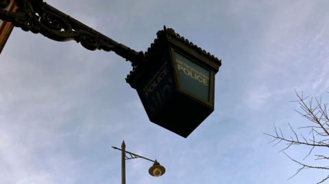 Met Police lantern against a blue sky with street lamp  and tree visible in the sky