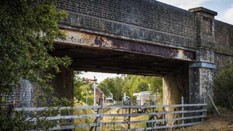 A brick railway bridge spanning a narrow passageway, with weathered steel beams visible beneath the structure. A wooden fence runs across the foreground, separating the viewer from the area under the bridge. Through the opening, several small buildings, a level-crossing gate, and a traditional railway semaphore signal can be seen in the distance. Trees, shrubs, and dry grass surround the scene.