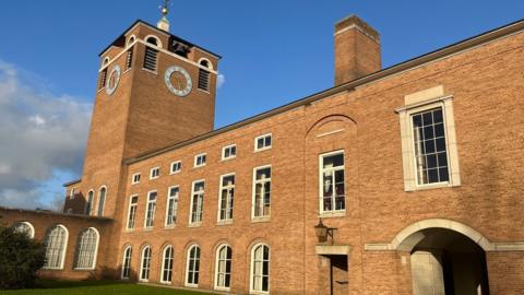 County Hall in Exeter. A large brick building with a tall clock tower featuring arched and rectangular windows, with white stone detailing around some of the frames. There is a prominent clock tower, with a golden weather vane at the top. 