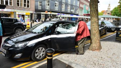 A black taxi is parked on the side of a busy road. The passenger door is open and a man wearing a red coat and black hat is about to get inside.