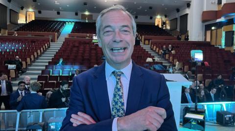 Nigel Farage smiling with an open mouth looking directly at the camera, wearing suit and tie and his arms crossed. He is standing in front of hundreds of auditorium seats, some of which are filled with people wearing suits and formal wear.