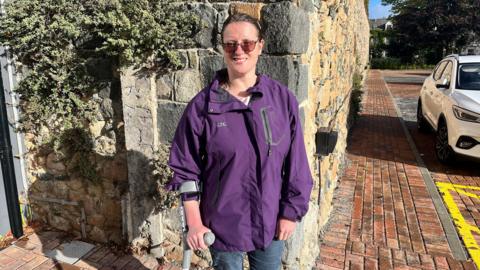 A woman in a purple jacket and light-sensitive glasses stands smiling in front of a wall, with her hand resting on a crutch.
