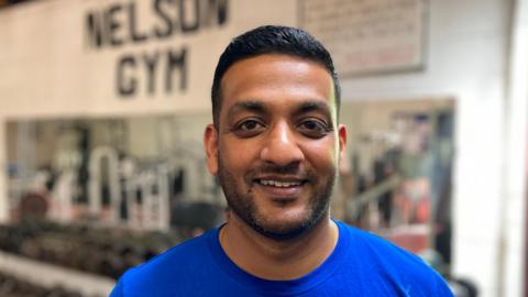 A man smiles toward the camera - he is wearing a blue T-shirt, behind him are dumbbells on a weights rack in a gym.