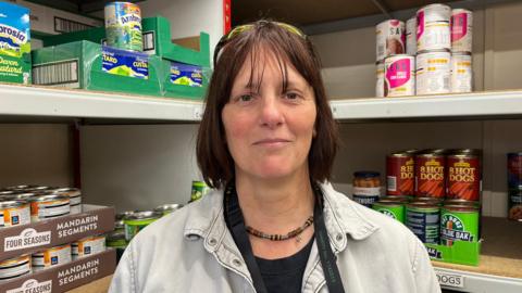 Liz looking into the camera with a neutral expression; she's wearing a grey denim shirt and a black and green Trussell Trust lanyard. She's stood in front of shelves stocked with supplies - you can see tins of mandarin segments, custard, hot dogs, and beans. 