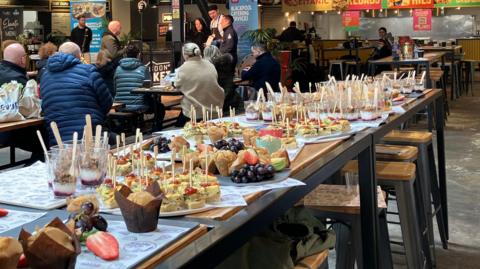 A table full of food in the foreground, each item has a wooden fork or spoon sticking out of the top. In the background, groups of people sit on tables looking towards a stage in far distance