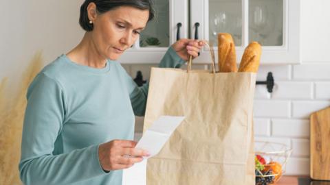 A woman looks at a receipt for her groceries. In her other hand she is holding a paper bag with two baguettes sticking out of the top