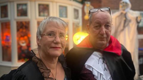 An elderly man and women pose together in black fancy dress costumes. She has short hair and glasses. He is wearing a dracula outfit. Behind them a house has been decorated with ghosts and pumpkins