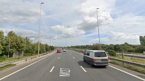 A screenshot from Google street view showing a three-lane motorway with cars driving along it. There are trees running down both sides, and it's a sunny day with some clouds in the sky.