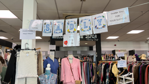 Interior of a charity shop with clothing rails full of dresses and jackets. A banner spelling "SADLY" hangs overhead alongside signs reading "We are closing," "Everything half price," and "Thank you for your support," indicating a closing-down sale.