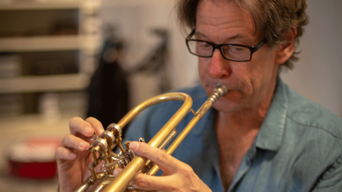 John Wallace playing a trumpet. He has the instrument to his mouth and is blowing on the mouthpiece. He is wearing black-rimmed glasses and has floppy hair. He is wearing a denim shirt. 