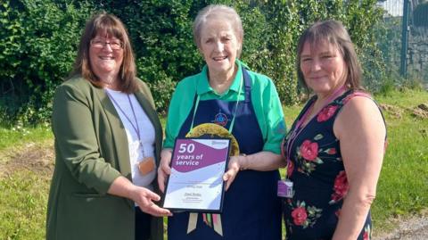 Dilys Brunt wearing a green top and navy blue apron, holding a framed certificate which reads "50 years of service". Two women stand either side of her smiling