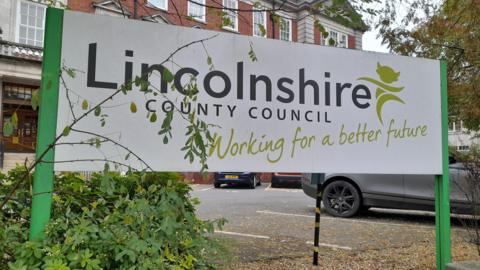 A close up of a sign saying Lincolnshire County Council, Working for a better future. It stands in front of a carpark and the council buildings.
