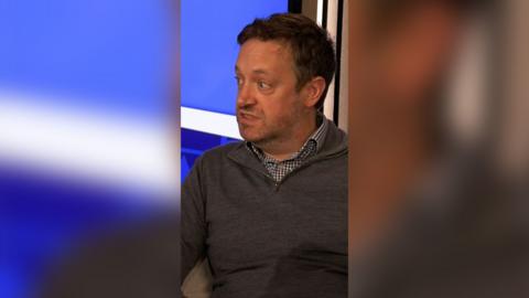 A picture of Alistair Murray in the Scotcast studio wearing a grey half zip jumper and black and white checked shirt underneath. He has short dark hair and is looking off to the left of the picture.